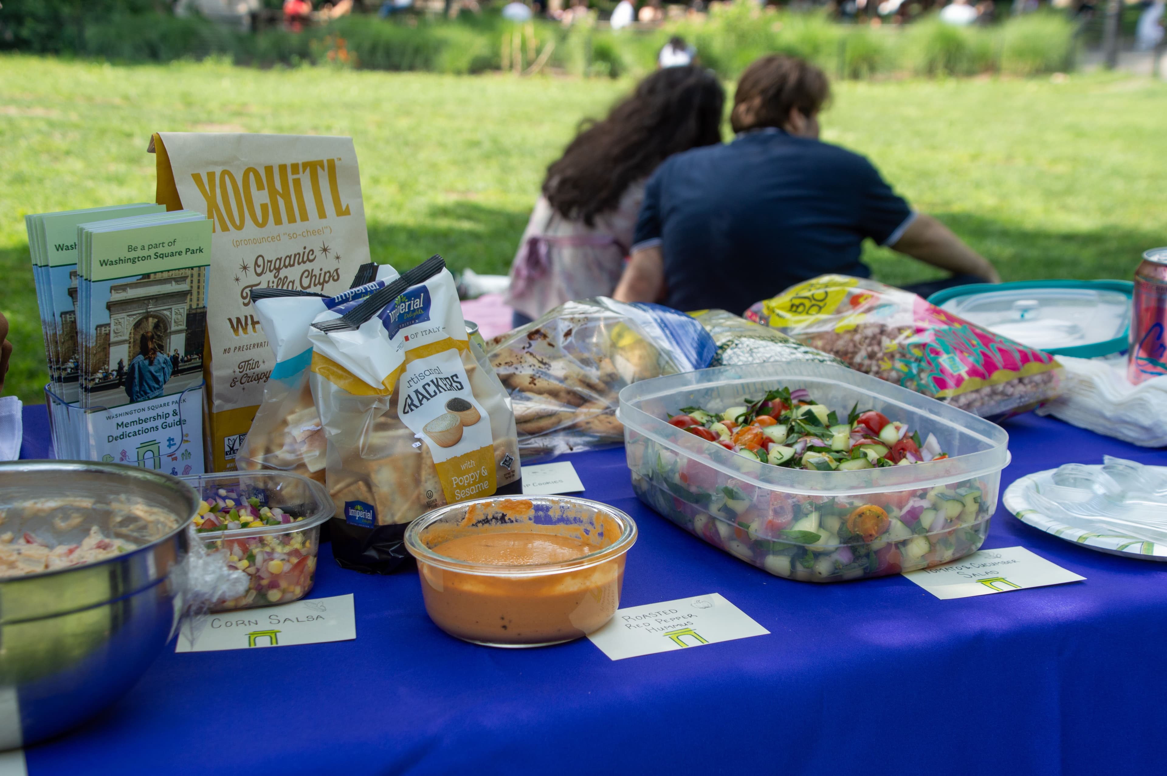 A spread of food, including hummus, salad, chips, and crackers, with labels that have the Washington Square Park Conservancy logo on them.